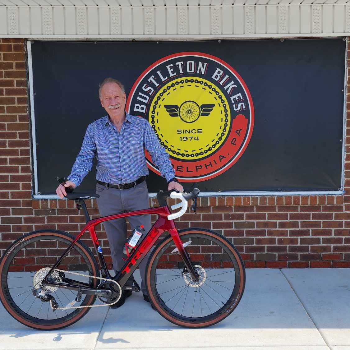 A smiling man standing outside in front of a Bustleton Bikes banner with a premium deep red carbon Trek road bike featuring tan wall tires