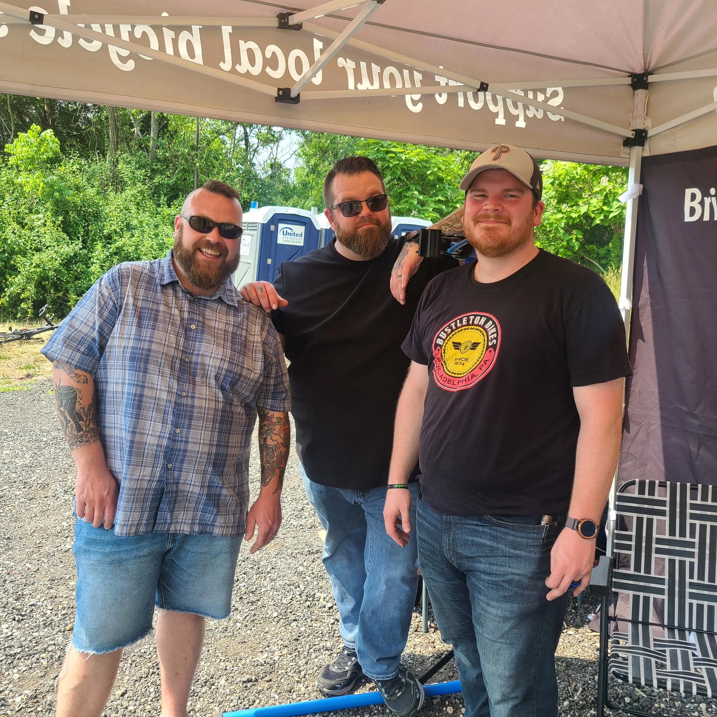 Three smiling men standing under an event tent at a rest stop during a community bike ride event.