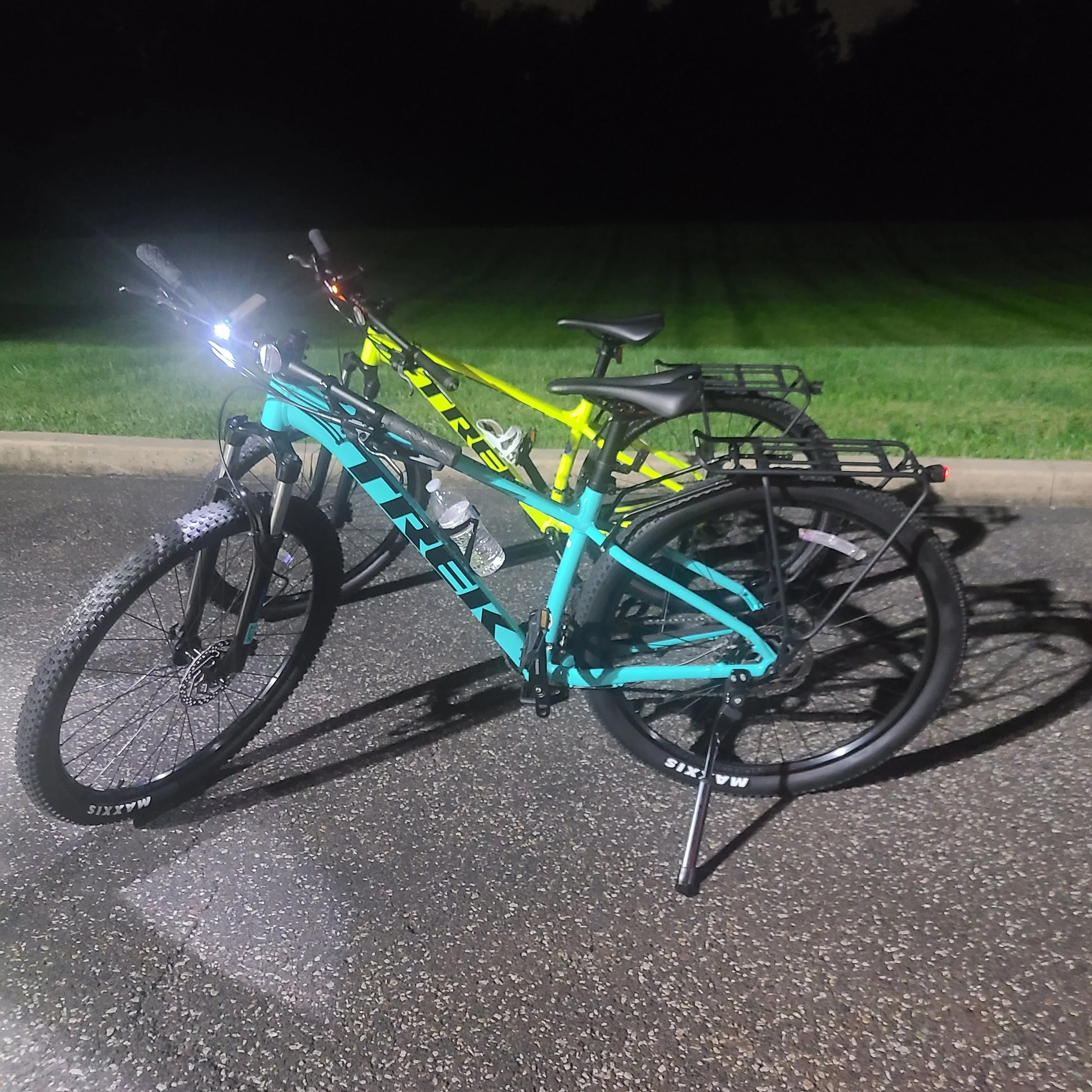 A turquoise Trek Marlin and a neon yellow Trek Marlin mountain bike parked on an asphalt lot at night with active LED headlights.