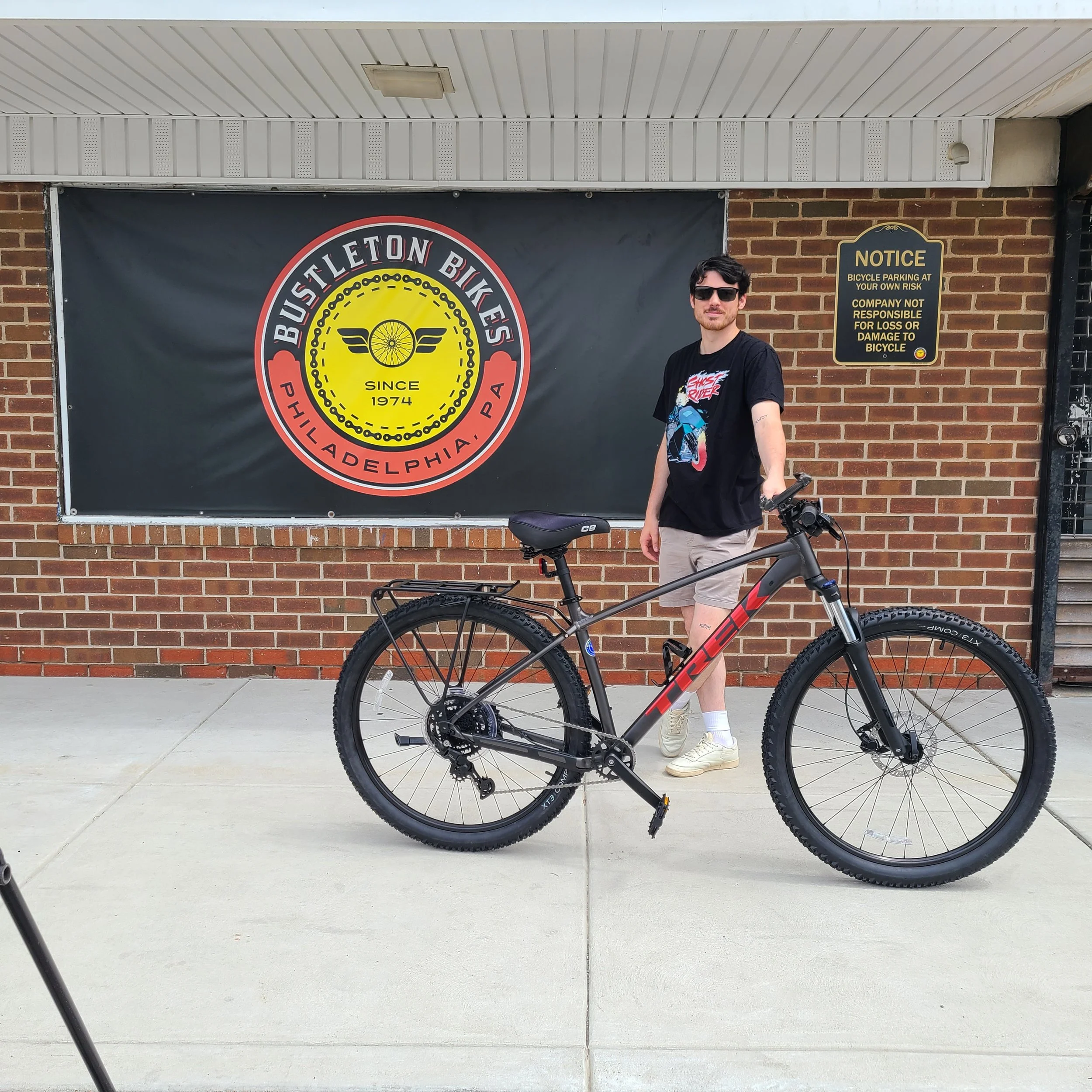 A man wearing sunglasses standing outside next to a dark grey Trek Marlin hardtail mountain bike outfitted with a rear utility rack.
