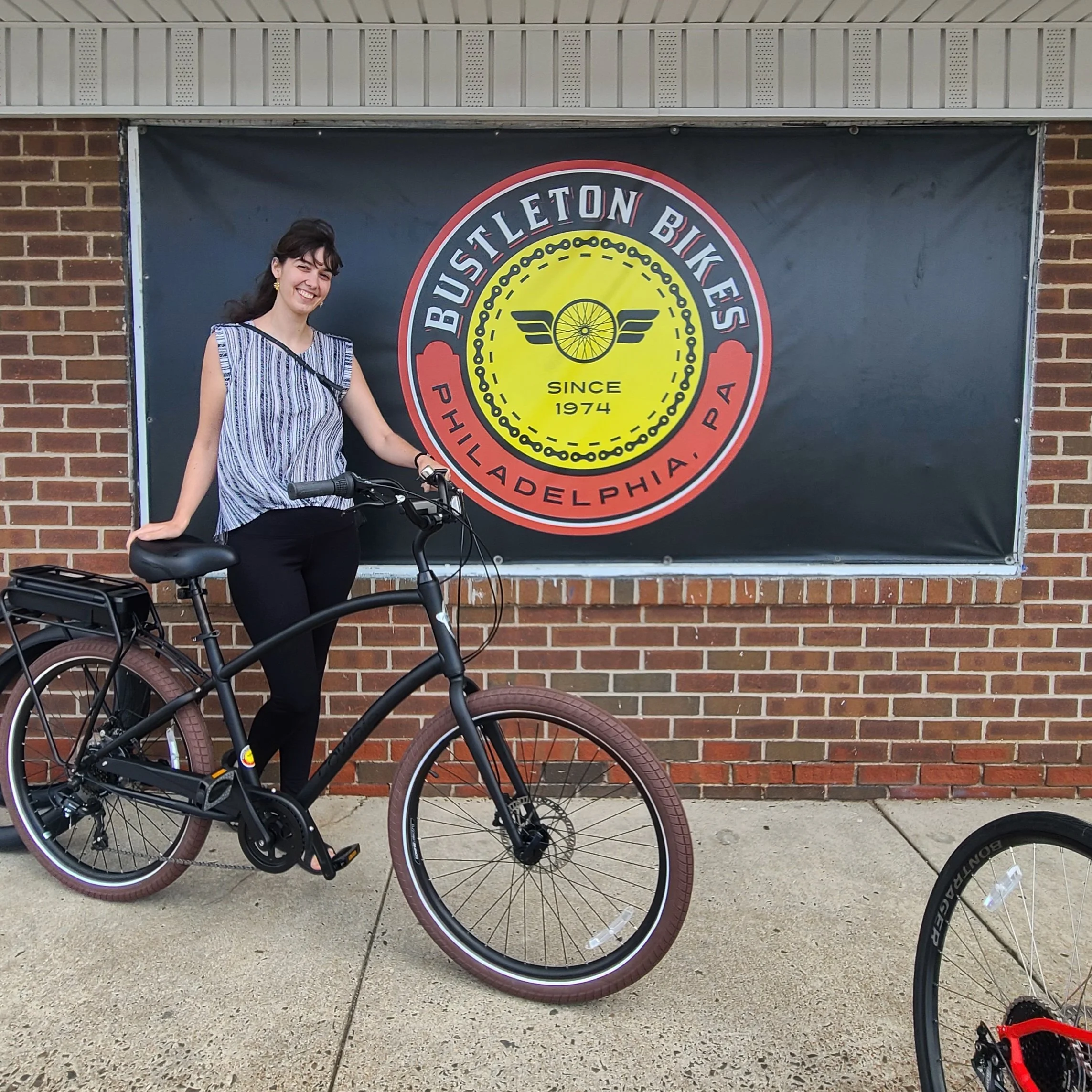 A smiling woman posing outside a bike shop with a matte black Electra Townie Go step-over electric cruiser bike with brown tires.