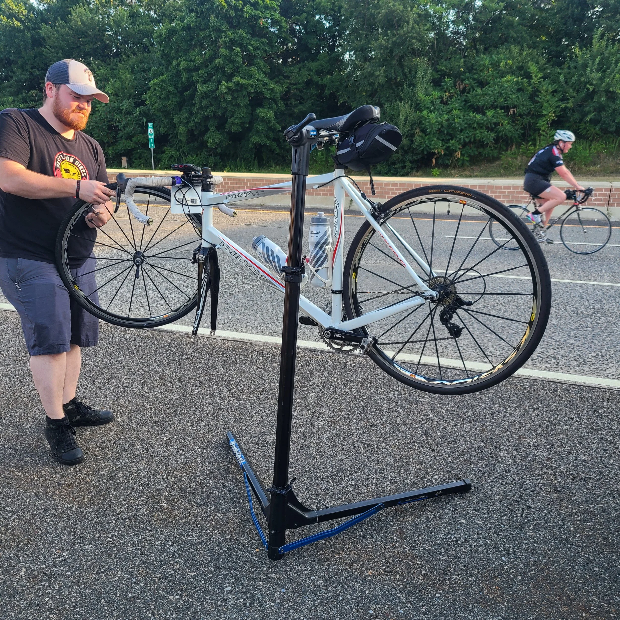 Jordan wearing a black Bustleton Bikes t-shirt services a white Ridley road bike mounted on a portable repair stand on the side of the road.
