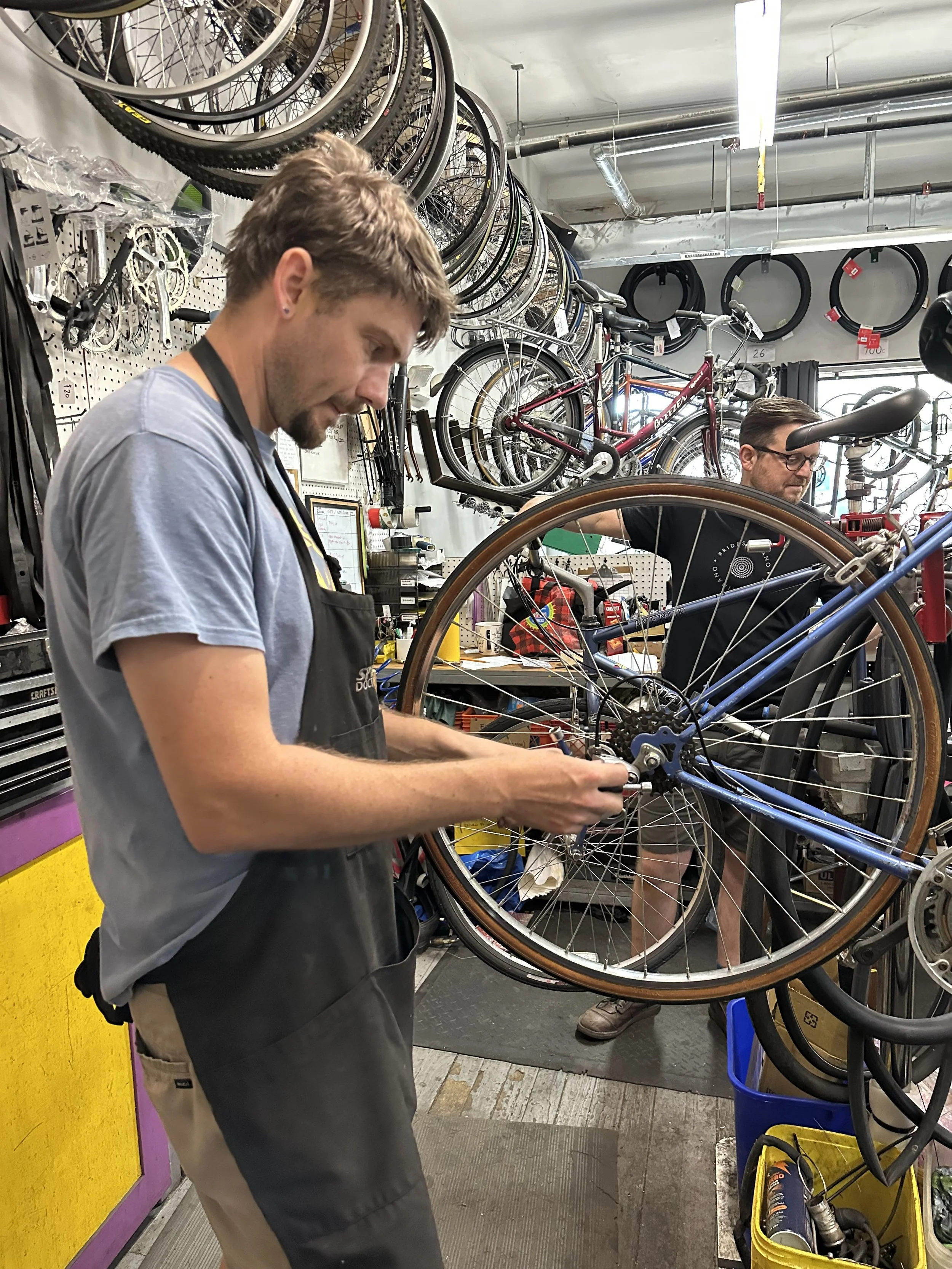 A bike mechanic in an apron tuning a rear wheel in the Neighborhood Bike Works community shop.