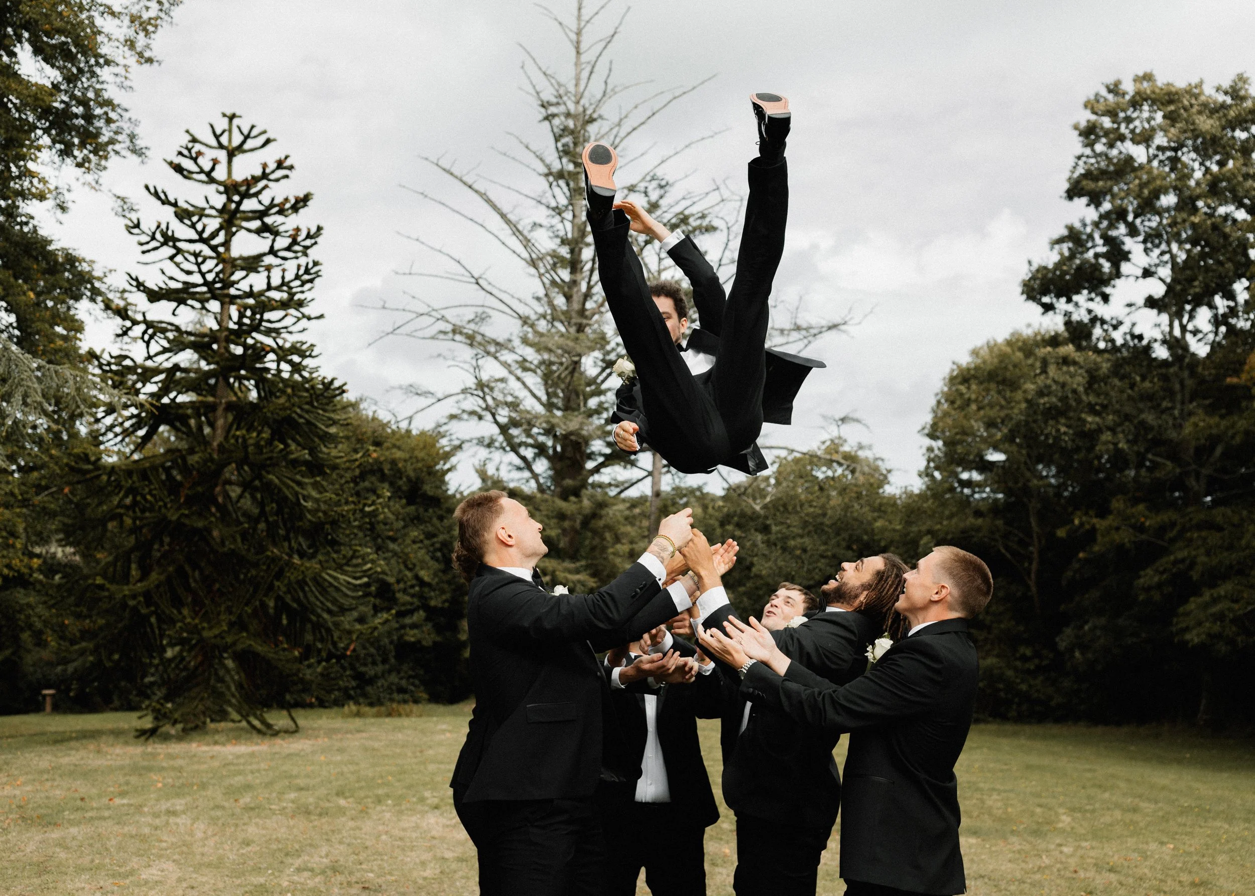 Groom in tuxedo being tossed into the air by group of friends in tuxedos outdoors during wedding celebration.