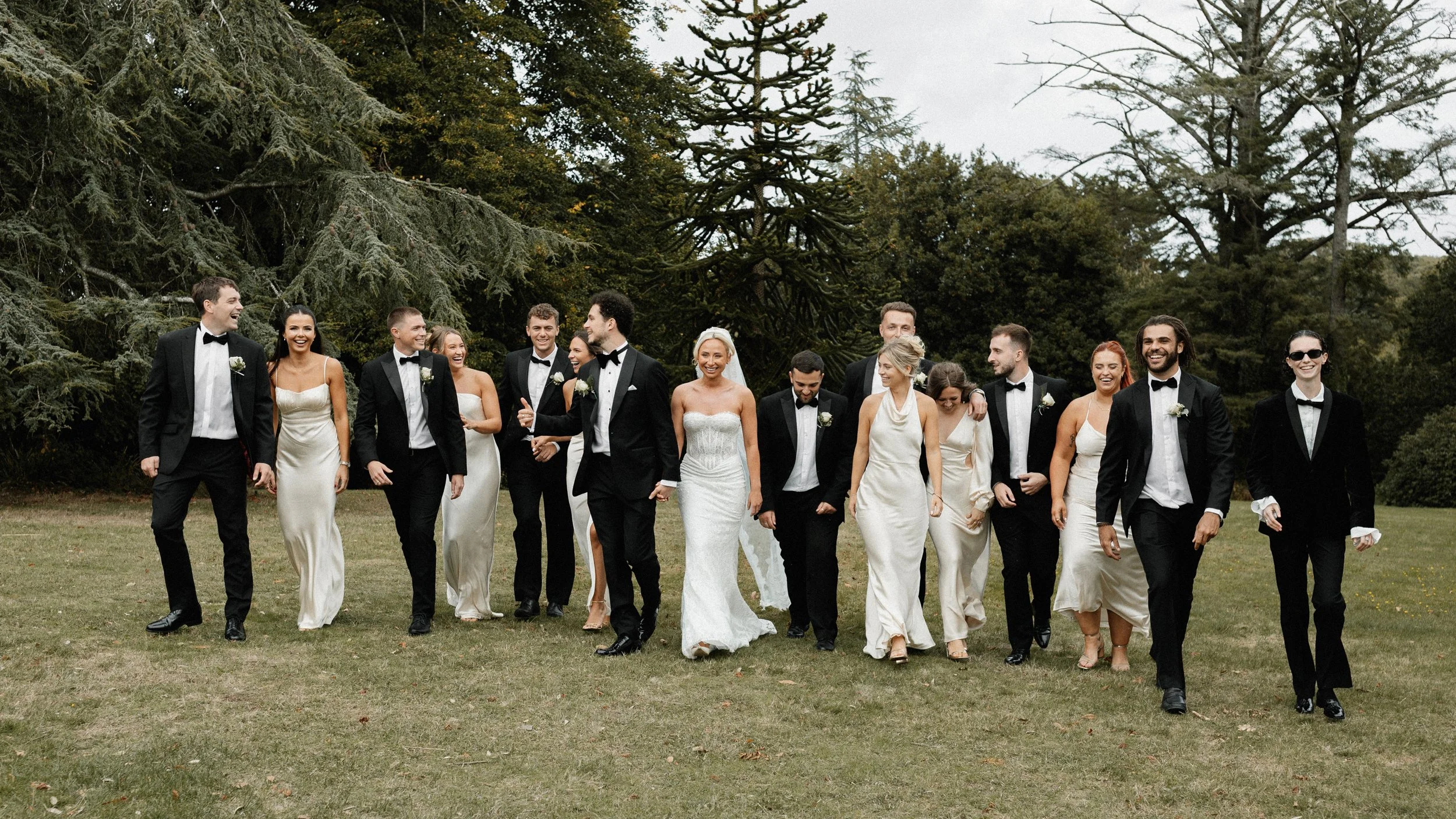 Group of wedding party members walking outdoors on grass, dressed in formal attire, with trees in the background, smiling and laughing.