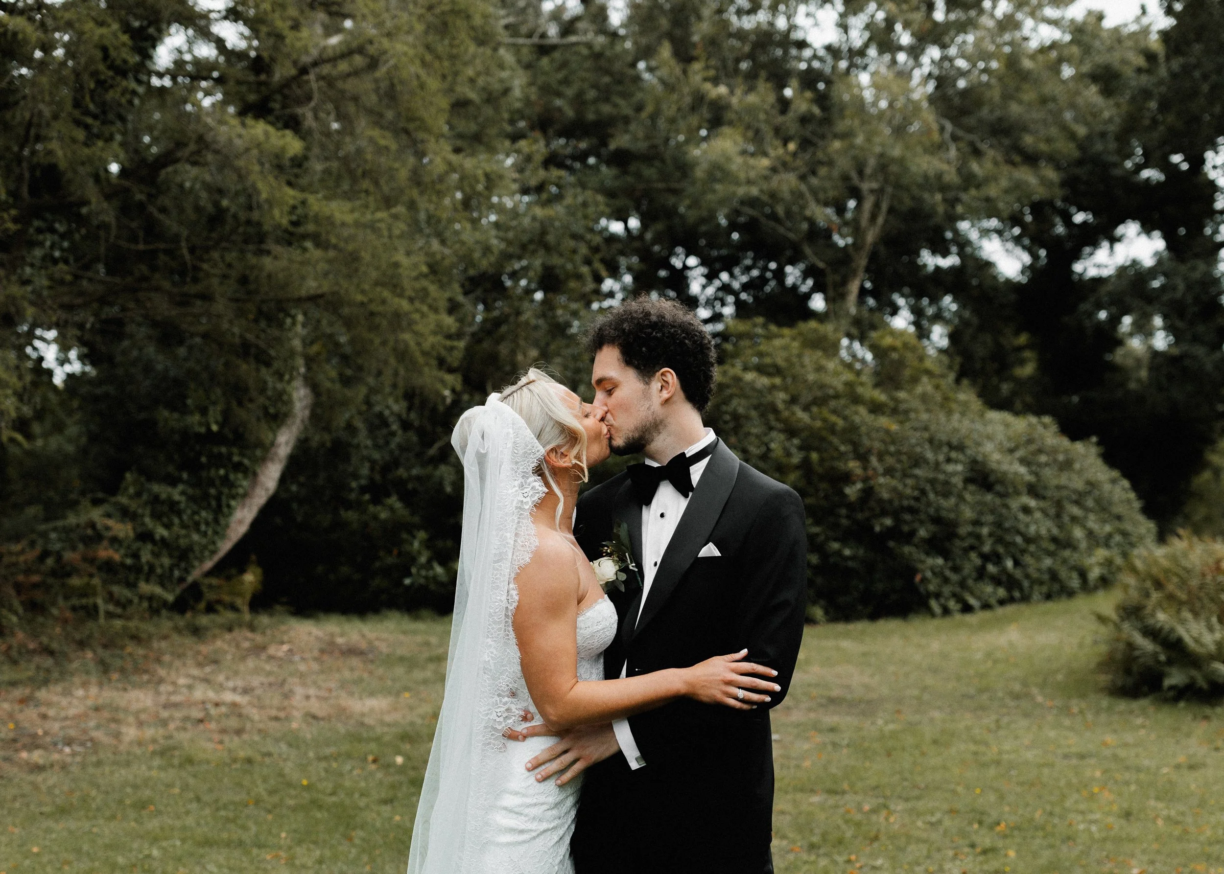 A bride and groom kiss in an outdoor setting surrounded by trees and greenery, with the bride wearing a white wedding dress and veil, and the groom in a black tuxedo with a bow tie.