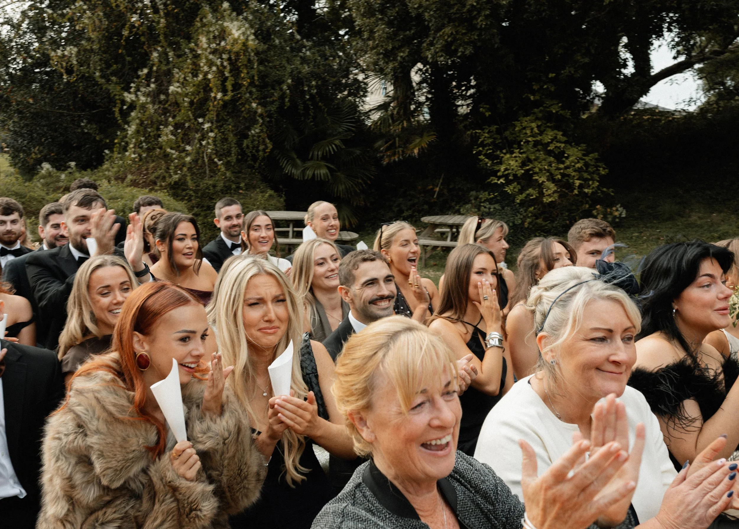 People attending an outdoor event, many are smiling and clapping, dressed in formal attire with some women in dresses and fur coats, trees and picnic tables are visible in the background.