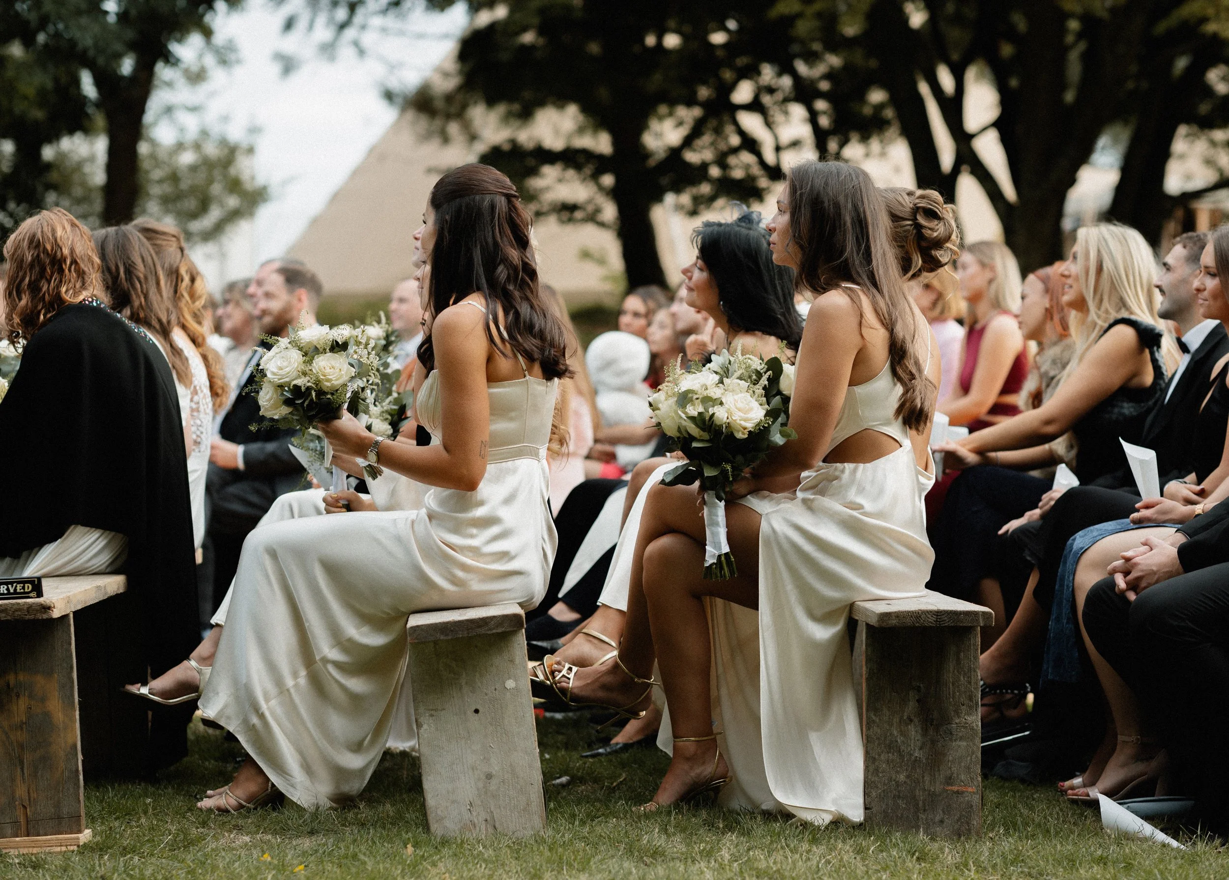 Women in cream dresses sitting on wooden benches during an outdoor wedding ceremony, holding bouquets of white flowers.