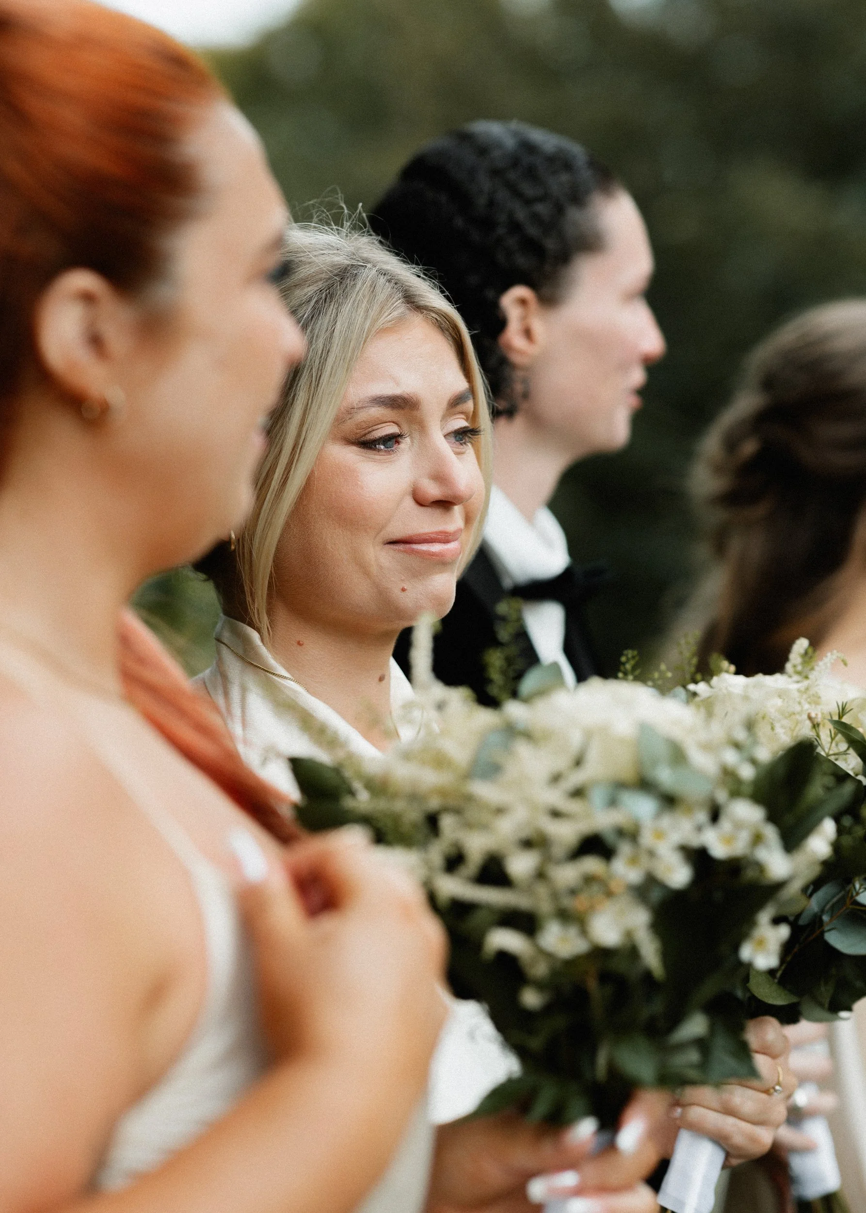 A woman with blonde hair and a serene expression holding a bouquet stands among a line of people at a wedding or ceremony outdoors.