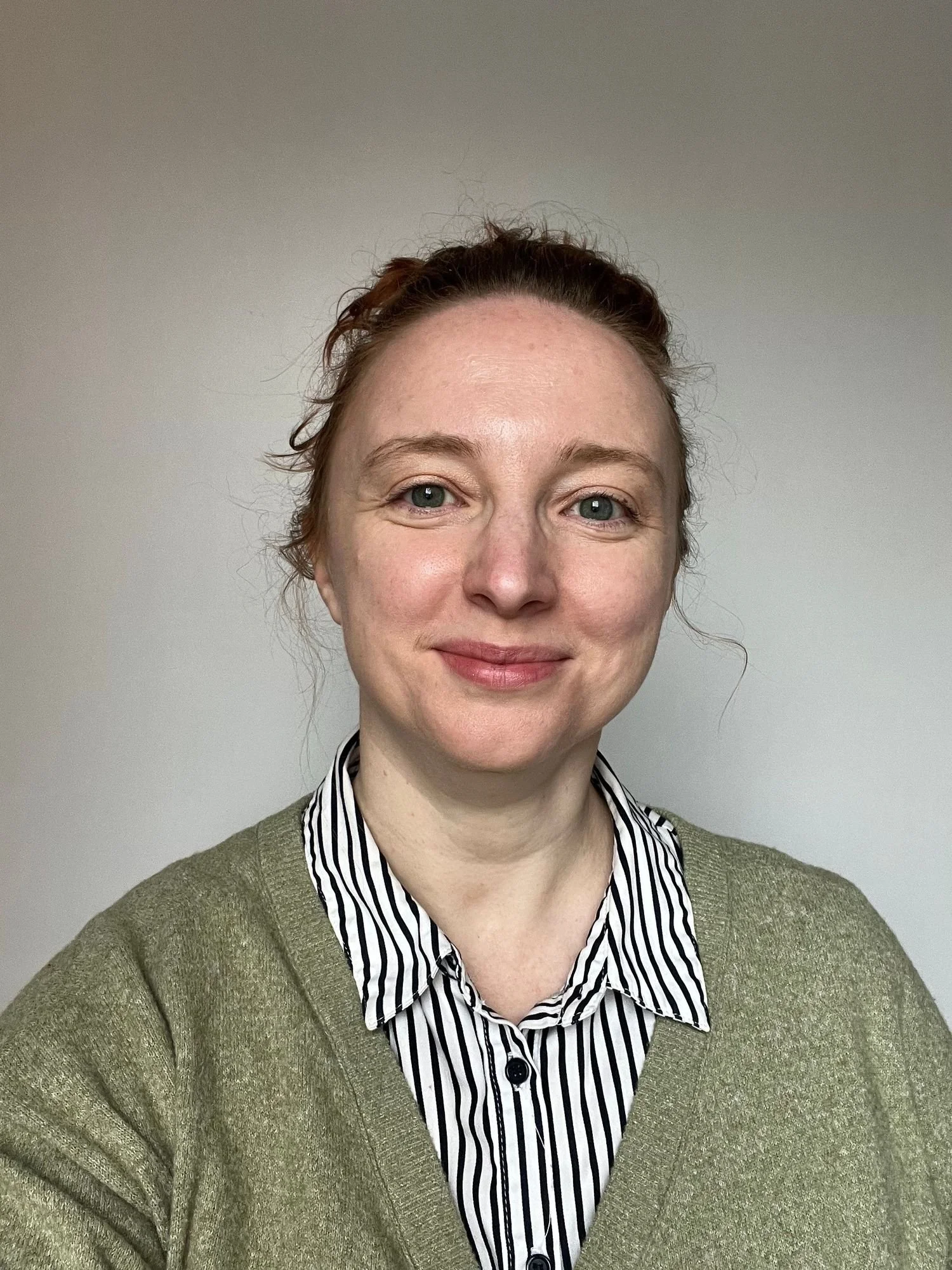 A woman with reddish hair tied up, fair skin, and blue eyes is smiling at the camera. She is wearing a striped collared shirt under an olive-green sweater, standing against a plain light gray background.