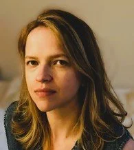 A portrait of a woman with shoulder-length brown hair, wearing a dark top, against a plain background.