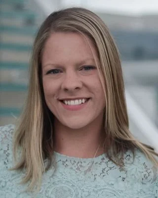 Portrait of a young woman with blonde hair smiling outdoors with a modern building in the background.
