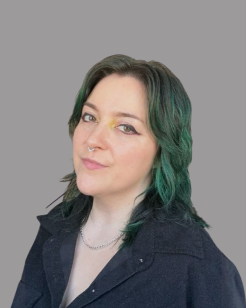 A woman with short, dark hair with green highlights, wearing a dark jacket and a necklace, looking at the camera against a plain gray background.