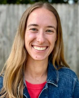 A young woman with long blonde hair smiling, wearing a denim jacket and a red shirt, standing outdoors with a wooden fence in the background.