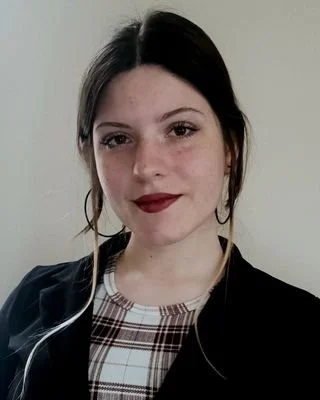 A young woman with dark hair, light skin, and red lipstick smiling at the camera, wearing a black jacket and a plaid top.