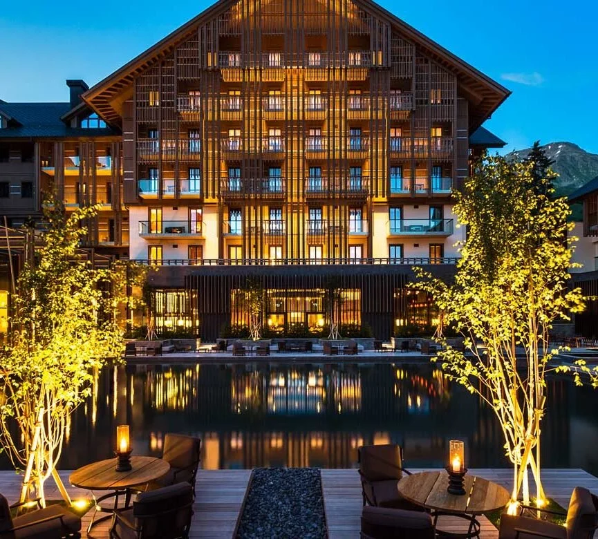 Luxury hotel at night with lit windows, outdoor seating with tables and chairs, illuminated trees, and a water feature reflecting the building, mountains in the background.