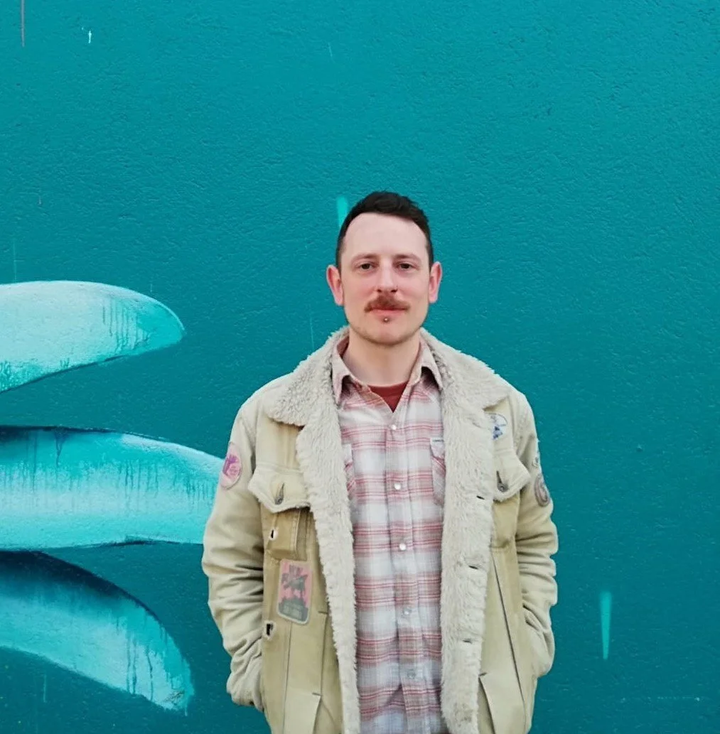 A man standing in front of a blue wall with abstract graffiti art. He has short dark hair, a mustache, and is wearing a beige coat with patches over a plaid shirt.