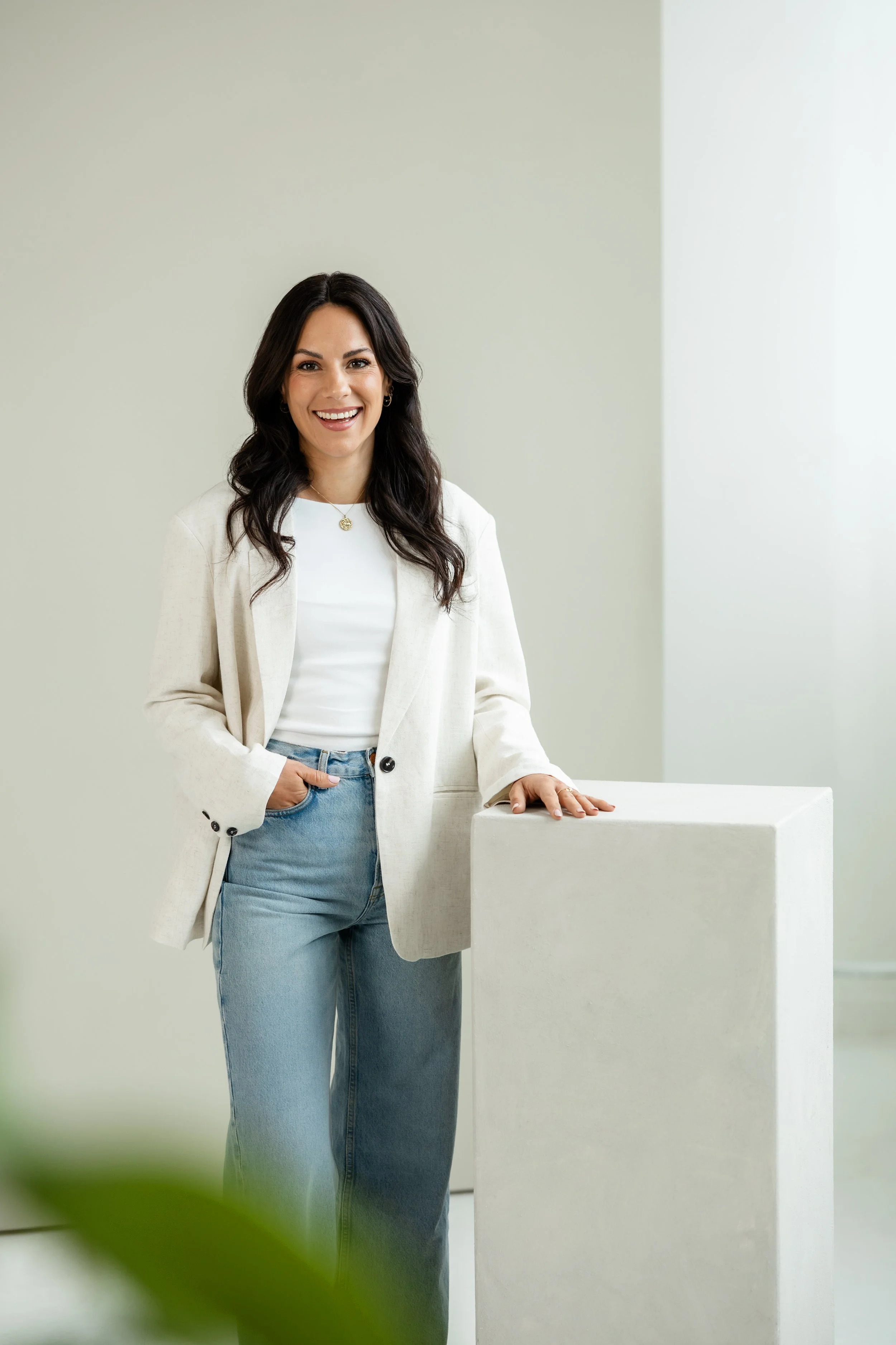 Eine lächelnde Frau mit dunklen, gewellten Haaren trägt einen weißen Blazer und blaue Jeans.