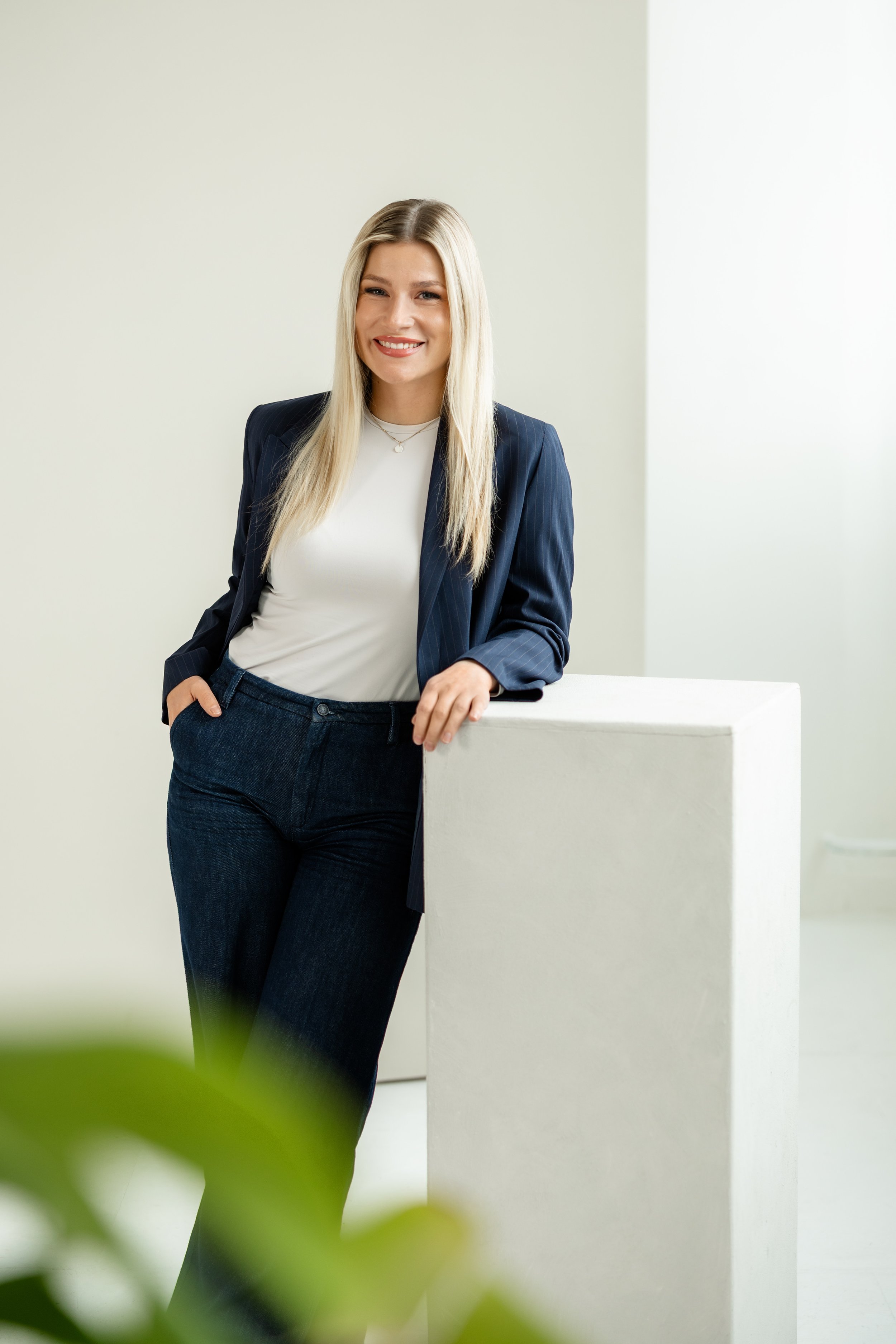 Junge Frau mit blonden Haaren in dunkler Businessjacke und weißen Hemd, die lächelnd an einer weißen Wand lehnt.