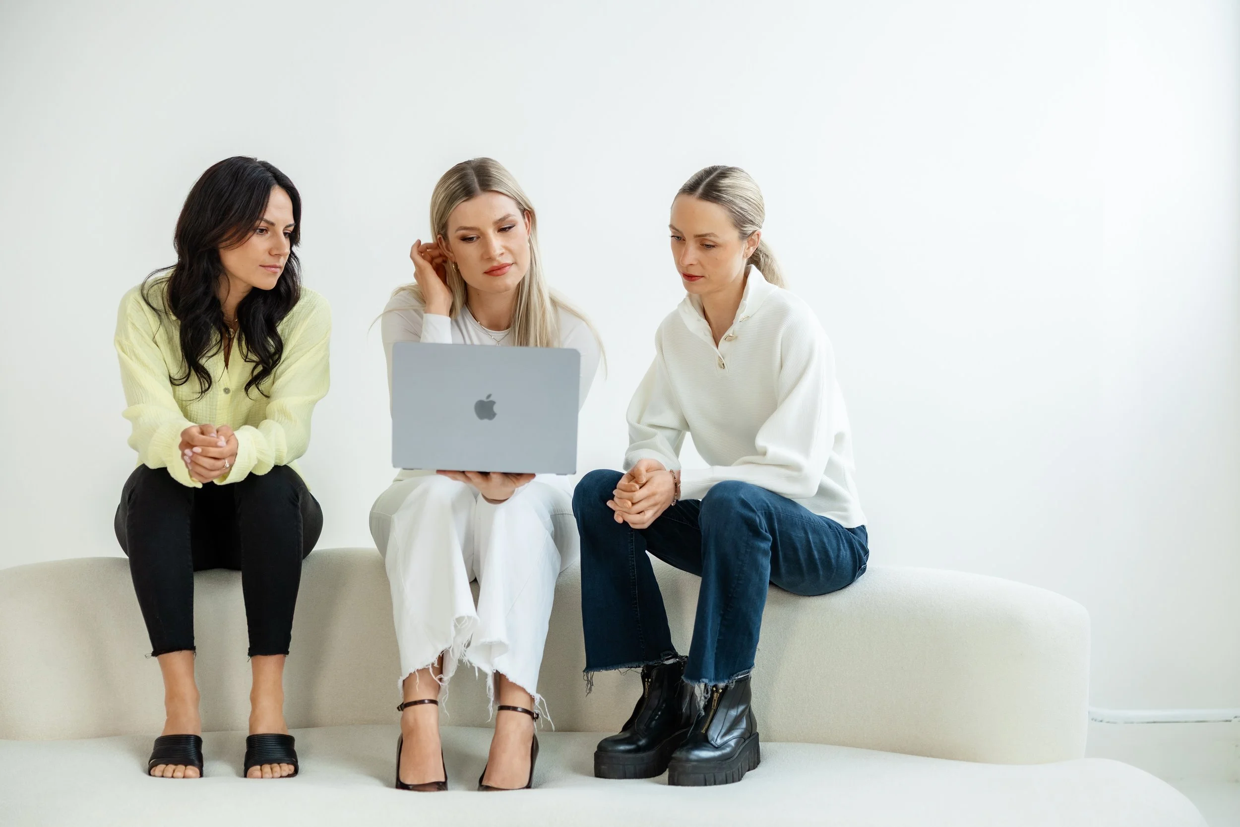 Drei Frauen sitzen auf einer weißen Couch und schauen auf einen Laptop, 2 tragen helle Kleidung, eine trägt dunkle Schuhe, eine trägt klobige schwarze Schuhe.