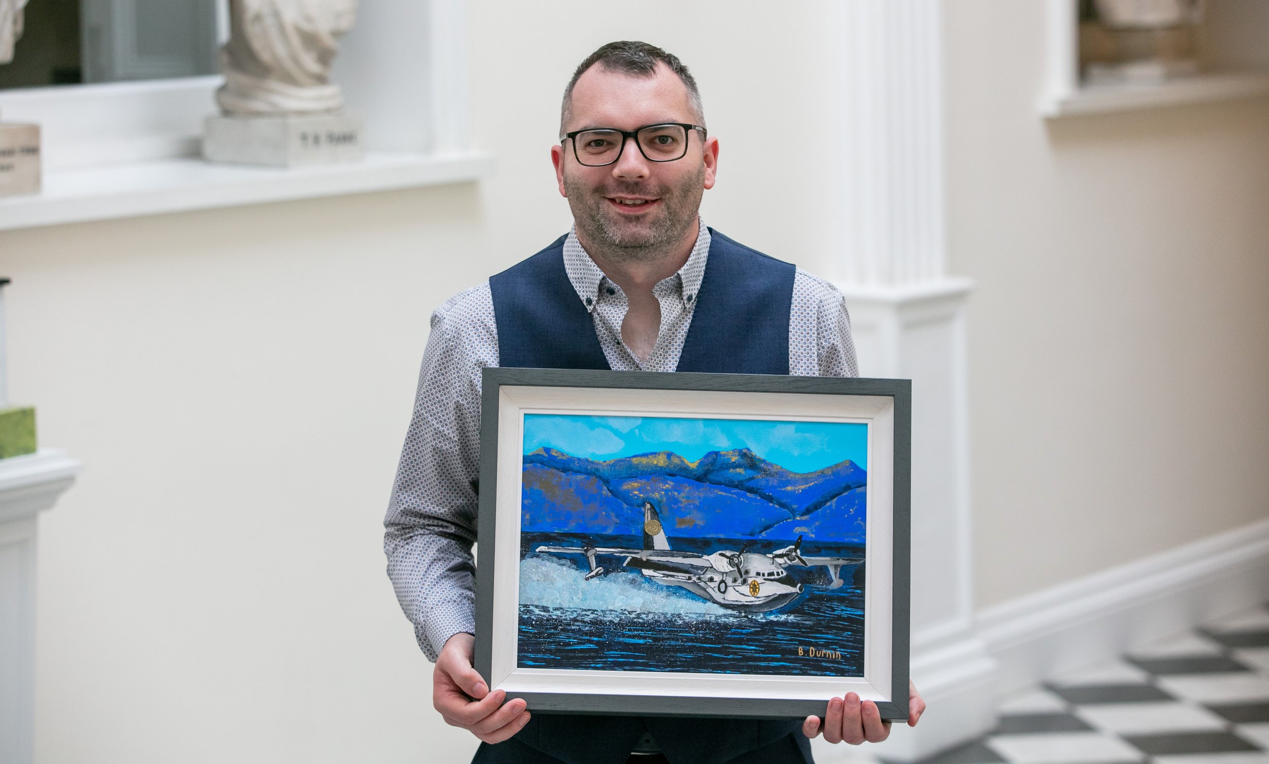 A man in a vest and patterned shirt holds a framed painting of a white seaplane on the water with mountains in the background.