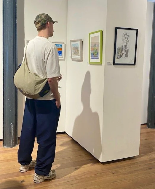 A person wearing a camouflage cap, a light-colored t-shirt, and navy sweatpants stands in an art gallery, looking at a series of framed artworks displayed on a white wall. The person carries a light-colored messenger bag over their shoulder and holds