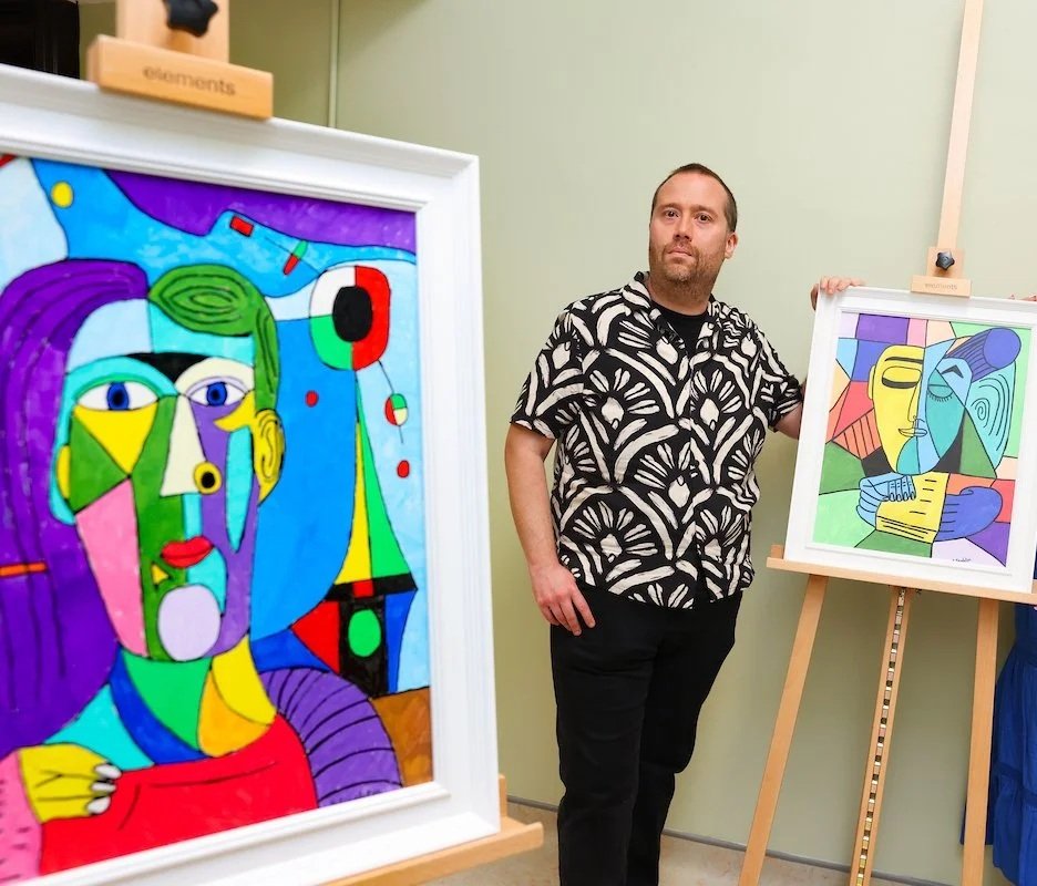 An individual stands beside colorful, cubist-style paintings displayed on easels and a frame.