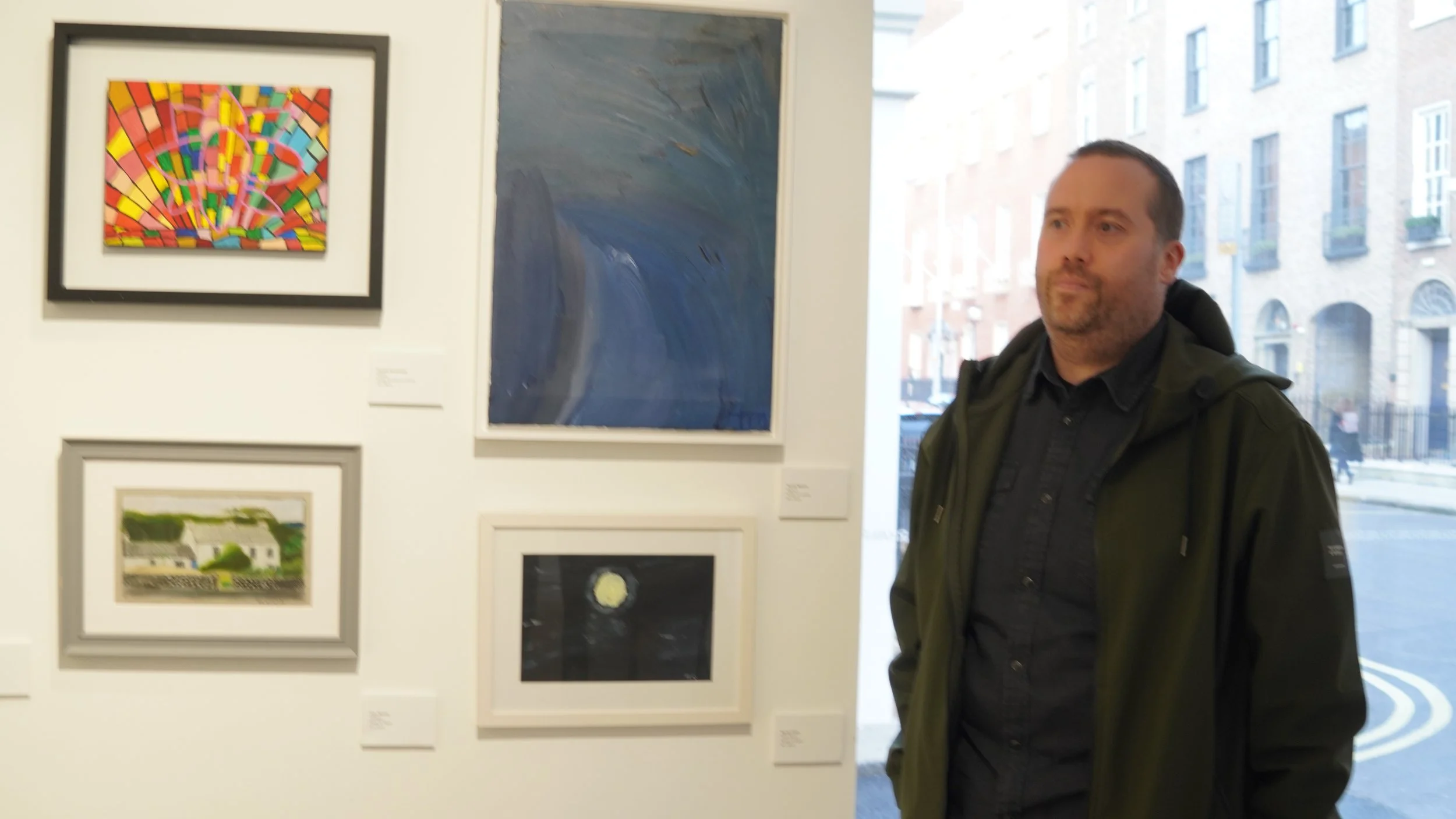 A man wearing a dark green jacket stands in an art gallery looking to his right. Behind him on a white wall are several framed artworks: a colorful abstract piece, a dark blue abstract painting, a watercolor landscape of a house, and a small dark pai
