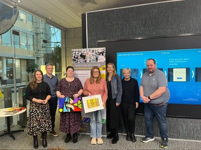 Group of seven people standing in front of a stand up banner. Two of them are holding colorful artwork. 