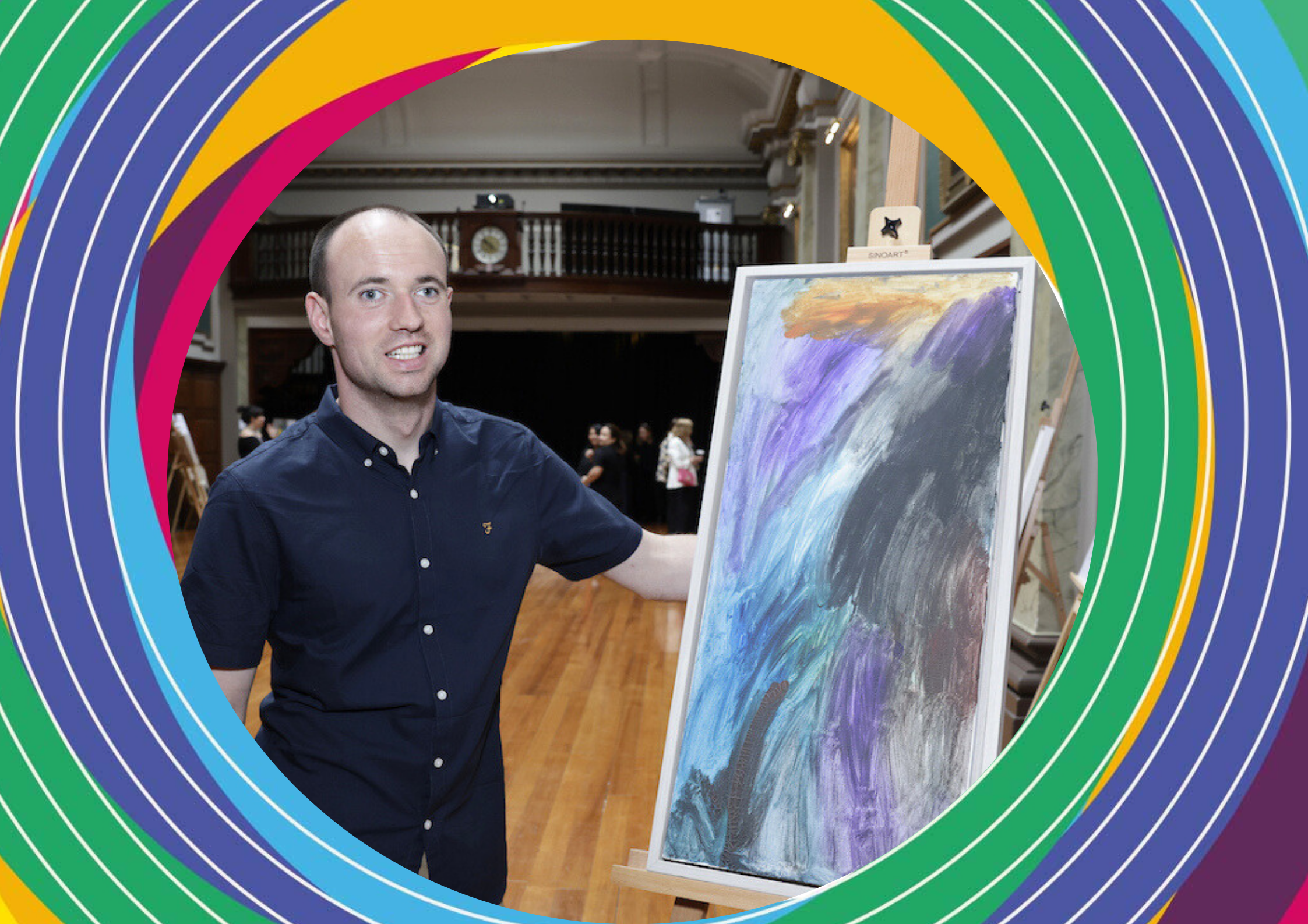Artist Tomás Malone, a man in a dark blue dress shirt, stands next to his abstract colourful painting titled 'Autumn'.