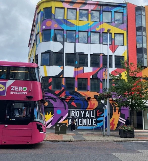 A bright pink double-decker bus passes a building covered in a vibrant, abstract mural featuring wavy lines in yellow, purple, blue, orange, and white, with "ROYAL AVENUE" written in large white letters on the lower section of the wall.