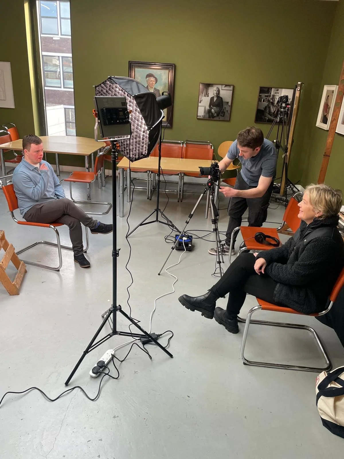Filming setup with a photographer adjusting a camera on a tripod, a person sitting on a chair in the foreground, and another person sitting on a chair in the background.