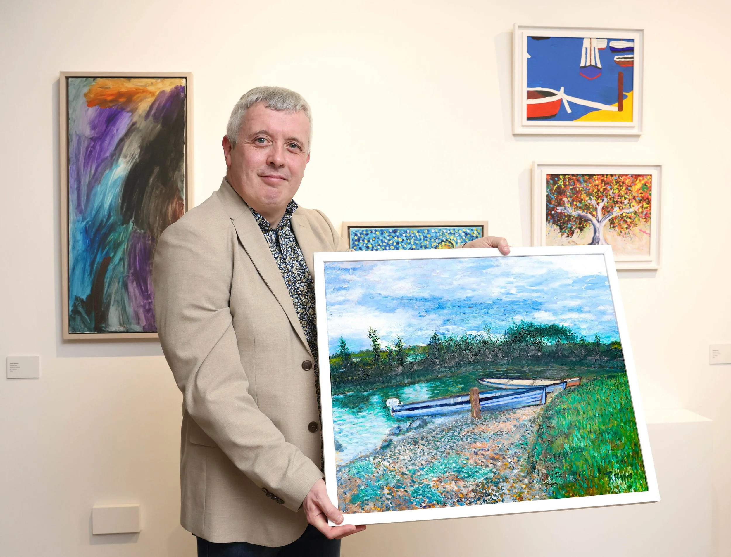 Man in a tan suit jacket holding up a framed painting of two blue boats docked on a pebble shore under a cloudy sky, with paintings on the gallery wall behind him.
