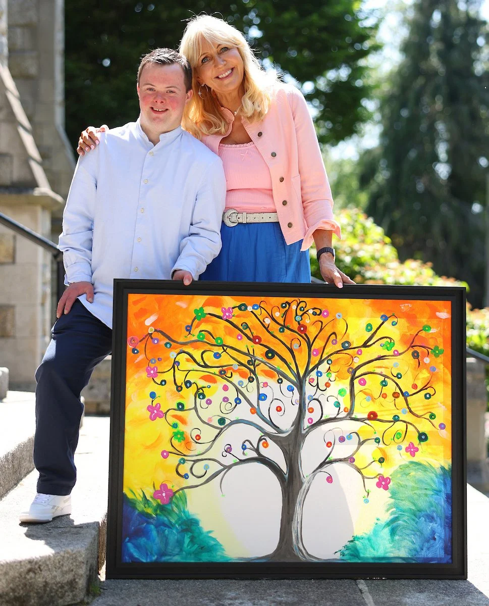 A smiling young man with Down syndrome and a smiling blonde woman stand outdoors behind a framed, colorful painting of a stylized tree with bright flowers and colorful berries against an orange and yellow background.