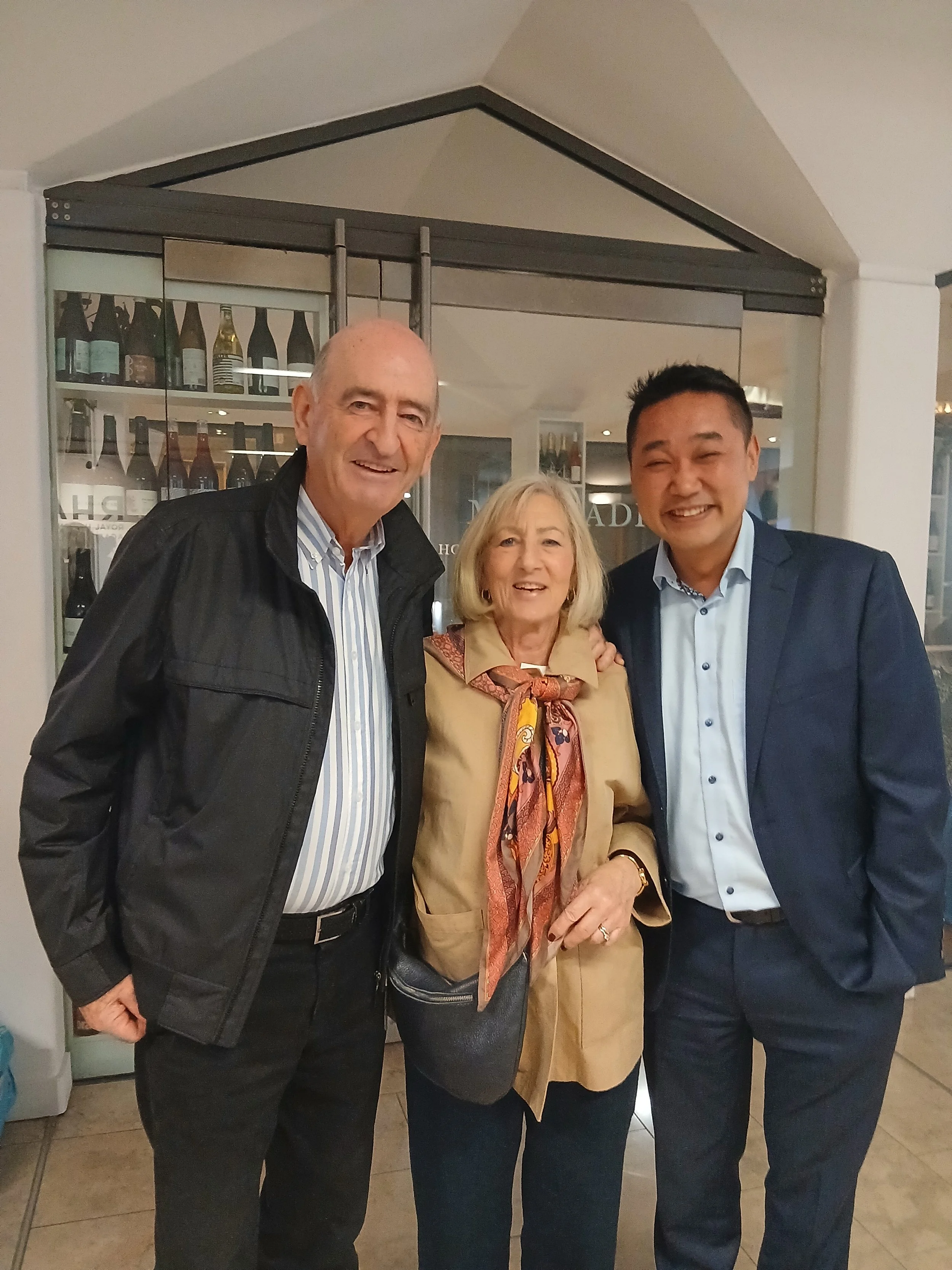 Three people, a man in a dark jacket, a woman in a tan jacket, and a man in a blue suit, pose together at the RHA opening