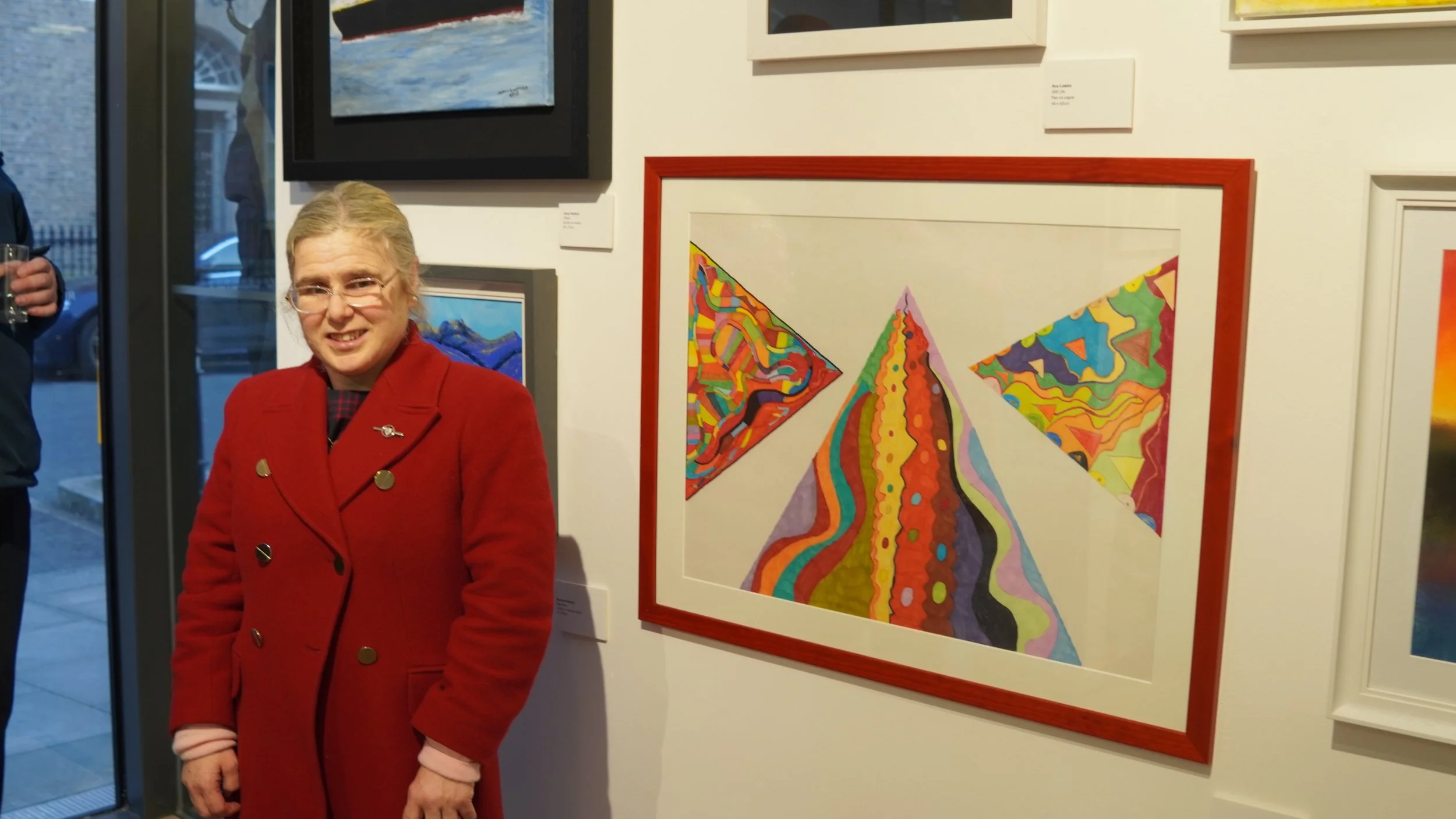 A person in a red coat stands in front of a wall displaying framed abstract artworks with colorful geometric and wavy patterns.