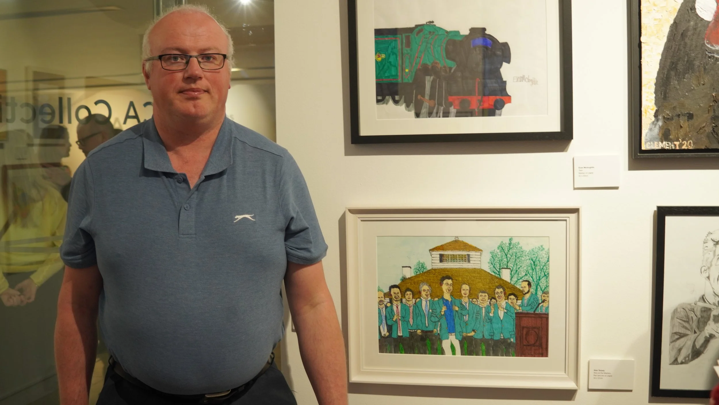 A man with glasses and a blue polo shirt stands in front of a wall displaying framed artwork, including a drawing of a train and a painting of a group of people.