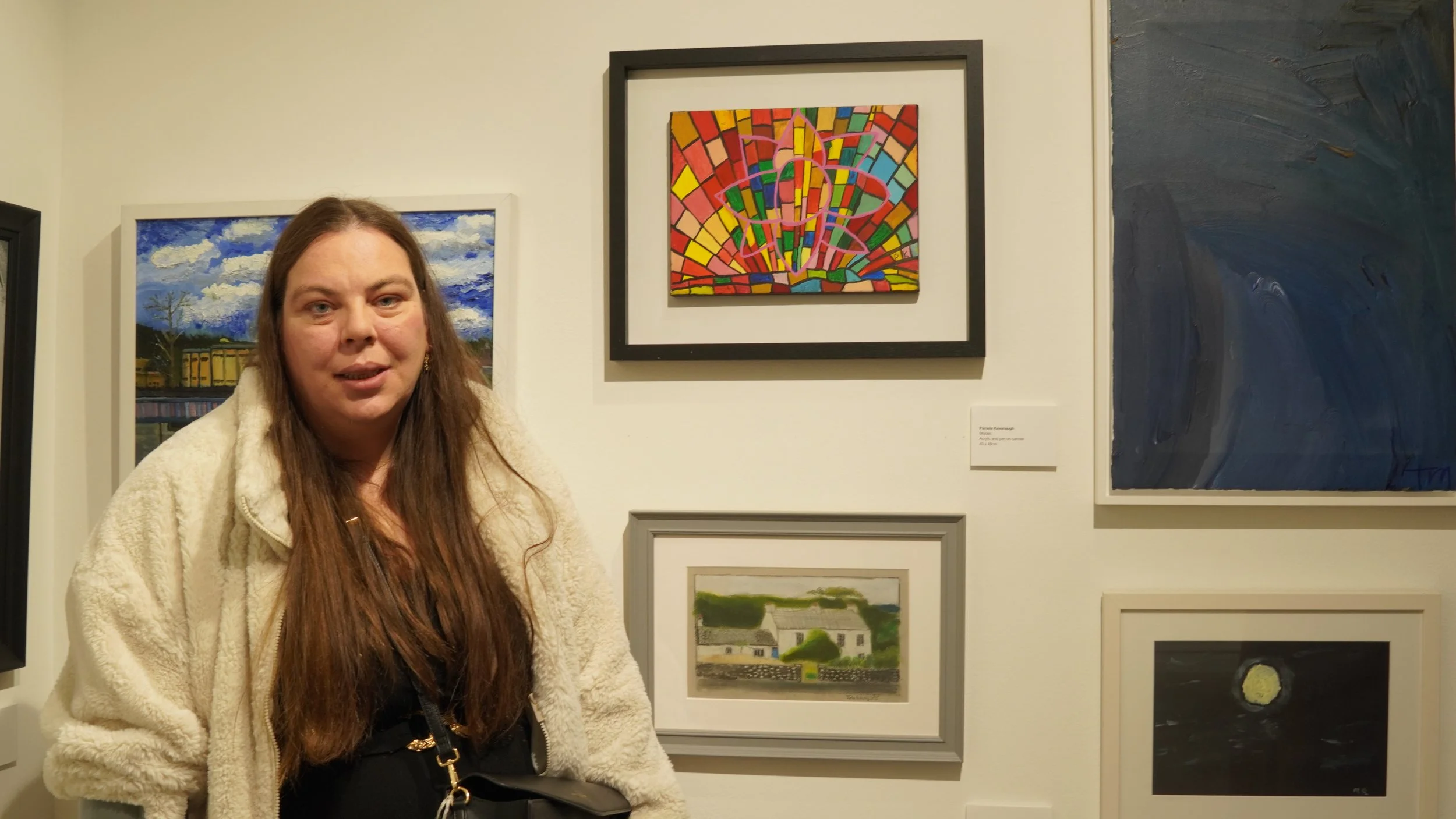 A man in a black puffer jacket and white polo shirt stands in front of a gallery wall featuring several framed artworks, gesturing towards a framed painting of a colorful sunset reflected in water.