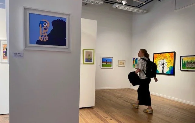 A person walks past artwork displayed on white walls in an art gallery, with a close-up of a blue and black painting on the left.