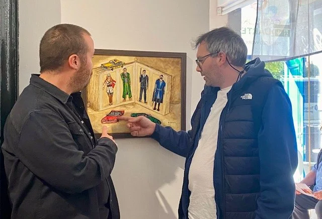 Two men stand in front of a framed painting that depicts a diorama with five figures, two toy cars, and a green object.