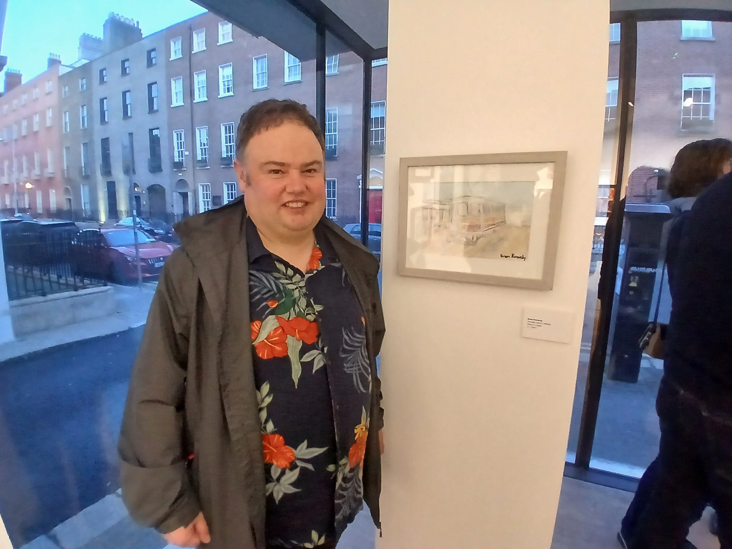 A man wearing a dark jacket over a colorful floral shirt stands in an art gallery, smiling at the camera. Behind him, a framed watercolor painting of buildings is displayed on a white wall. Through a large window, the exterior of a street with townho