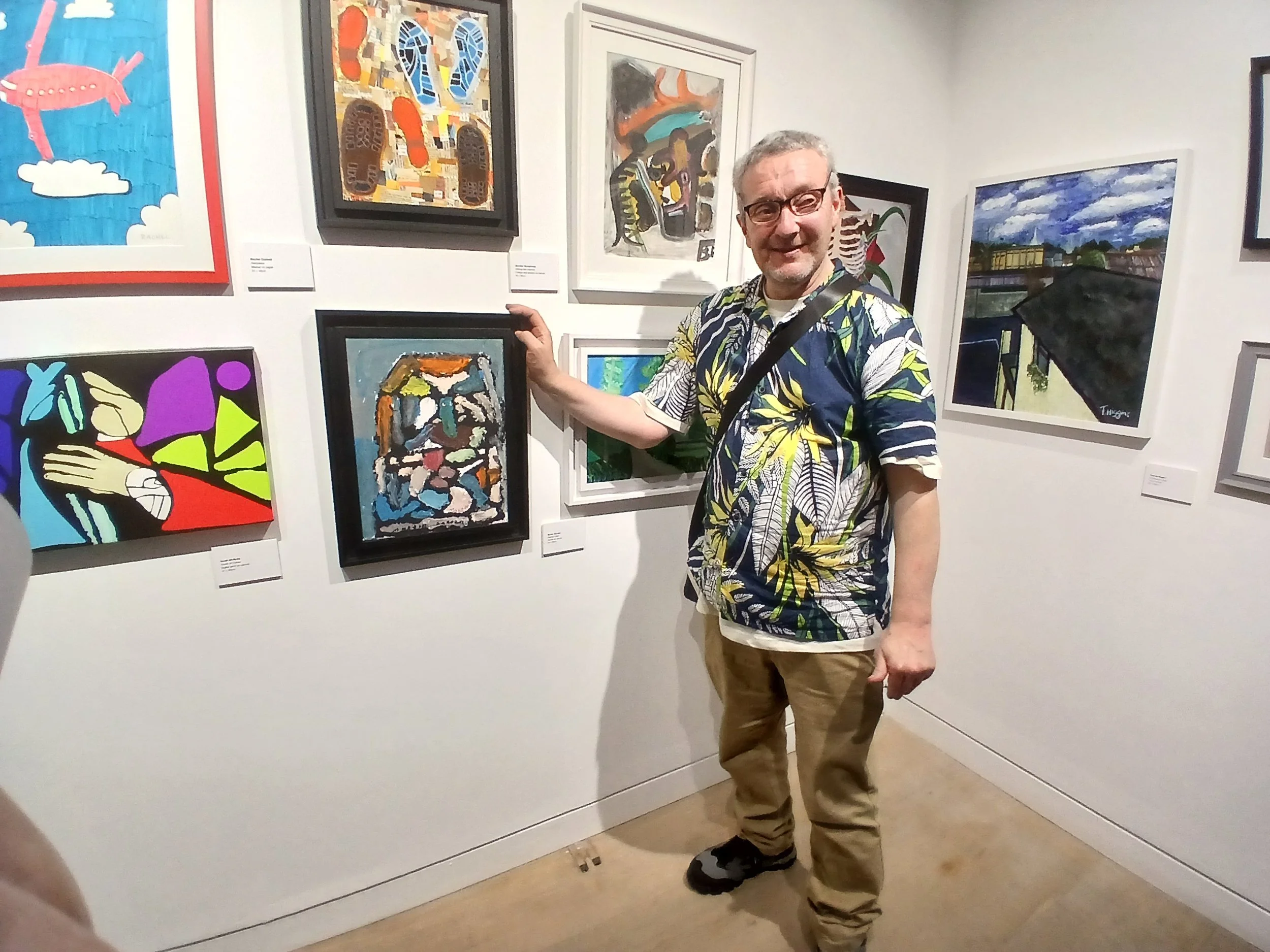 A man in a floral shirt stands in an art gallery, pointing to a framed abstract painting. Several other artworks are displayed on the white walls around him.