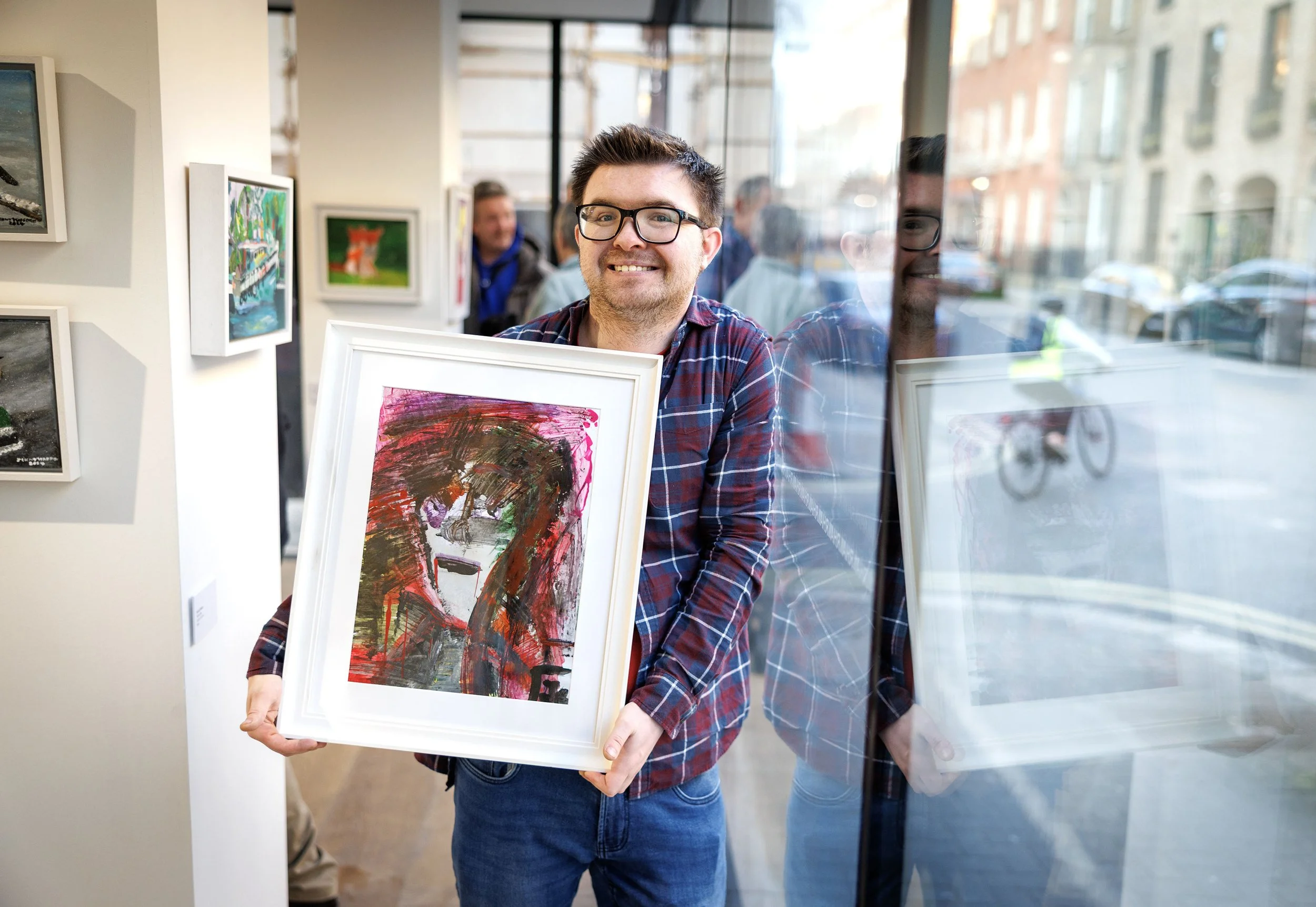 Man in plaid shirt and glasses smiling, holding a framed abstract painting in an art gallery, reflecting in the glass window behind him with a street scene visible.