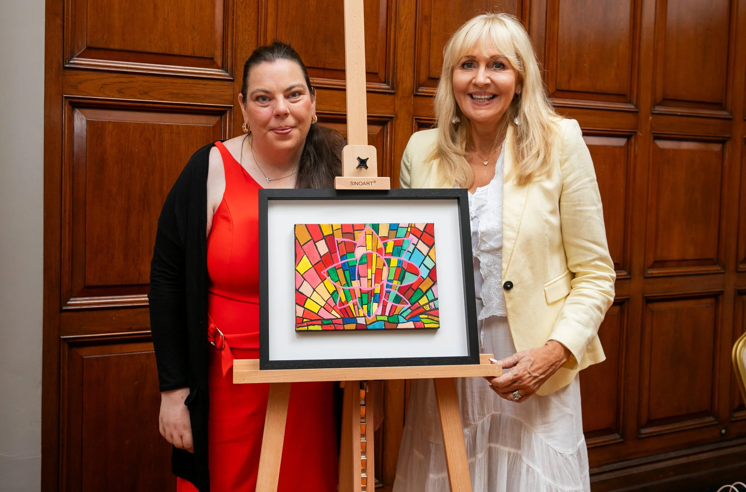 Two individuals stand beside an easel displaying a framed, colorful abstract painting of geometric shapes and vibrant lines.