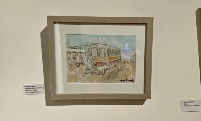 A framed pencil drawing of a vintage electric streetcar with yellow and red stripes, parked on tracks, with a blurred background and a signature at the bottom right.