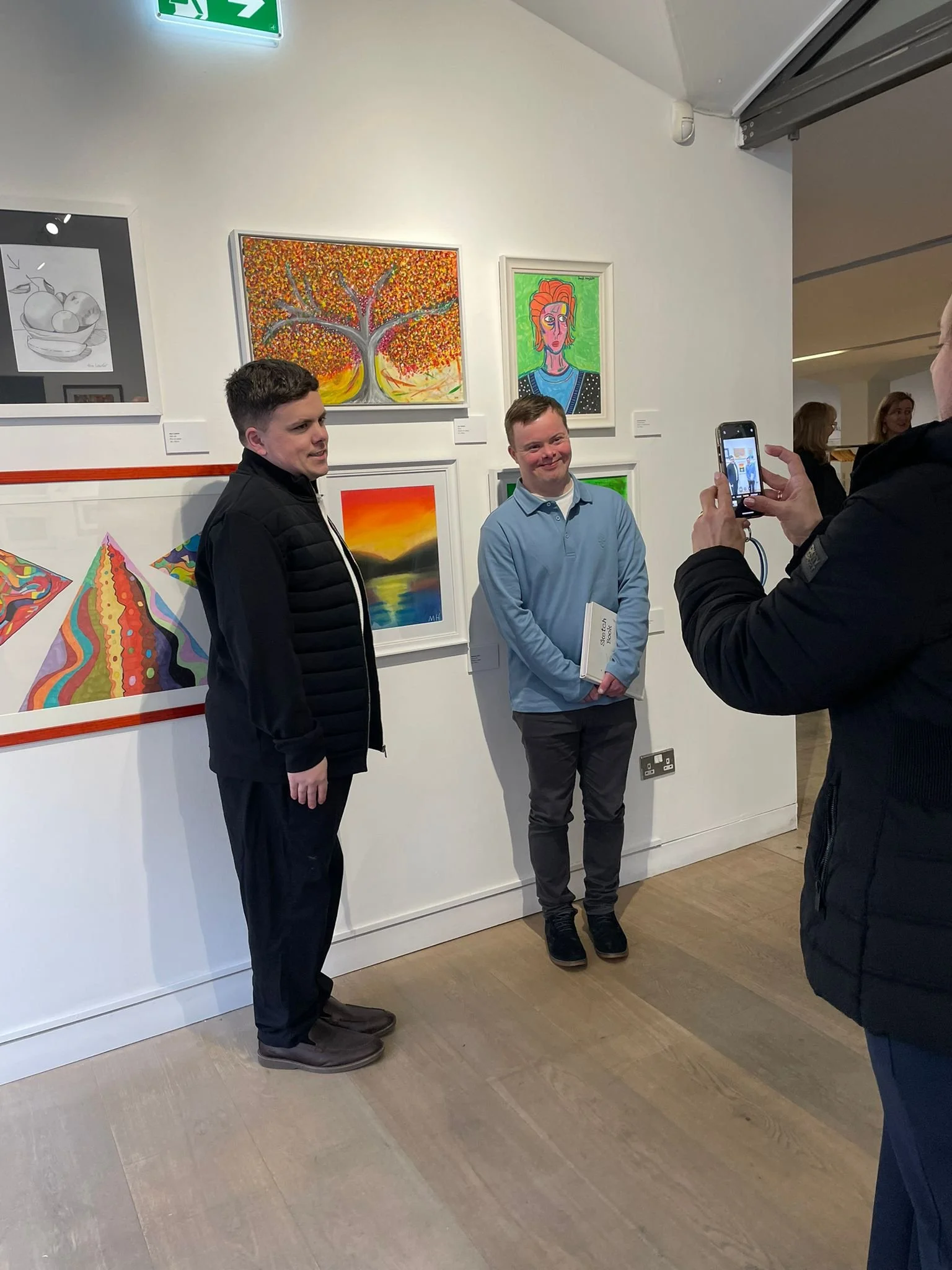 Four people stand in a brightly lit gallery space. In the foreground, a woman in a black t-shirt with a white logo and a man in a colorful, tropical-print shirt stand next to each other. To their right, another man in a similar tropical-print shirt a