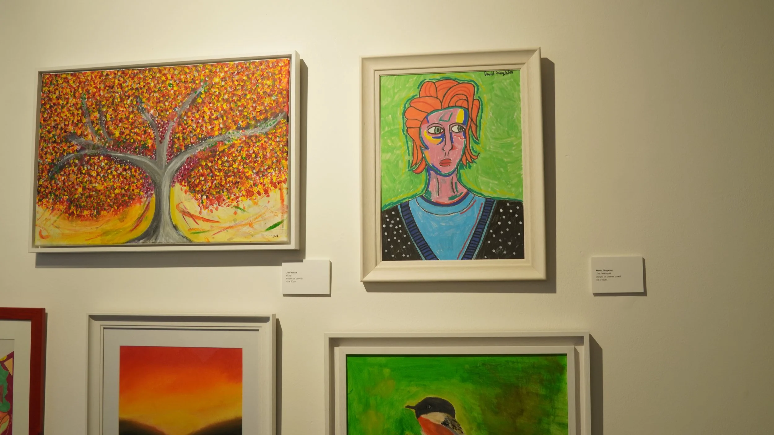 Framed artworks displayed on a white wall, featuring a vibrant abstract tree with autumnal foliage and a colorful, stylized portrait of a person with red hair.