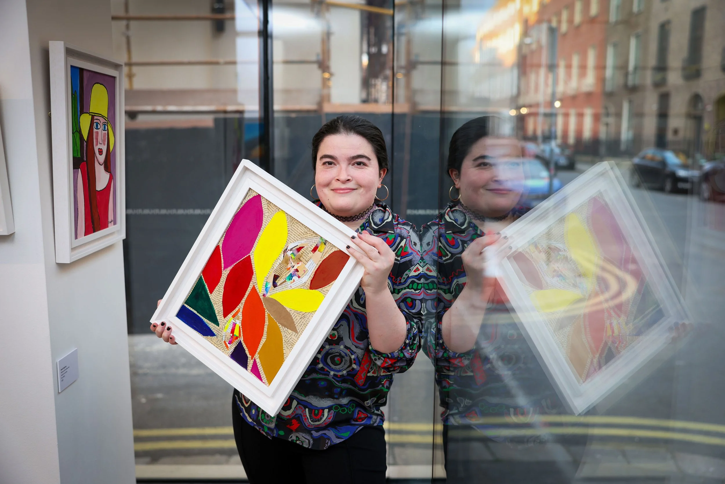 A person stands in an art gallery, holding a framed piece of art composed of colorful fabric petals arranged in a floral pattern on a textured background.