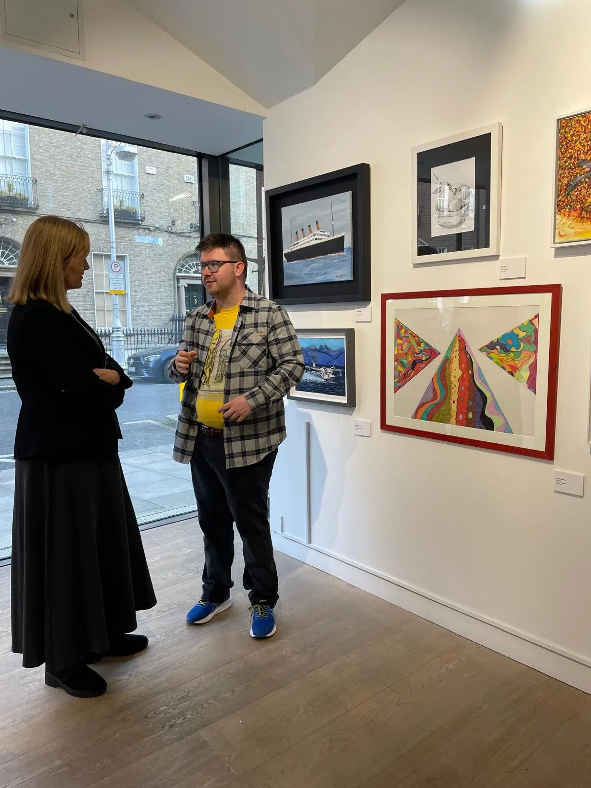 Two people stand in an art gallery, looking at paintings on the wall, with a large window to their left showing an urban street scene.