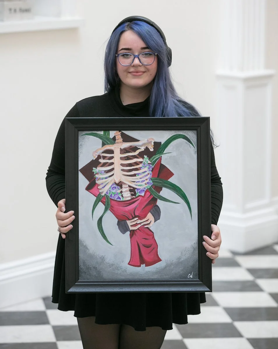 A person with blue hair holds a framed painting depicting a human rib cage intertwined with a bouquet of red flowers and green leaves, on a grey background.